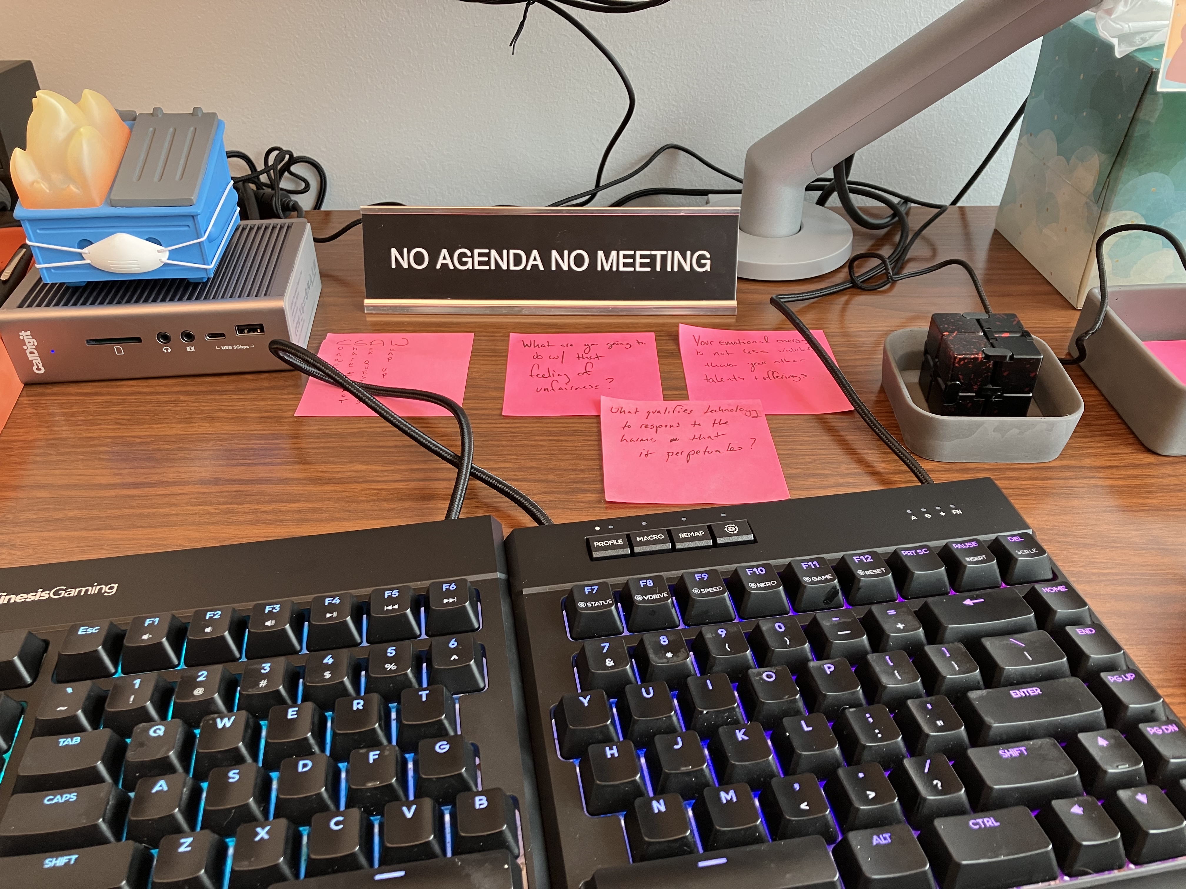 The top of a desk, with a keyboard taking much of the frame. At the back is a plaque that says 'no agenda, no meeting', next to a fidget cube, a cute plastic dumpster fire figurine, and a series of post it notes.